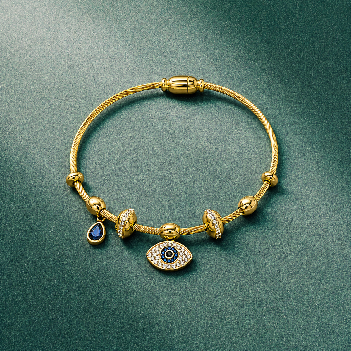 A gold cable bracelet with evil eye charms, featuring a central evil eye charm with blue and white details, a smaller teardrop blue charm, and textured gold spacer beads, displayed on a dark green textured background.