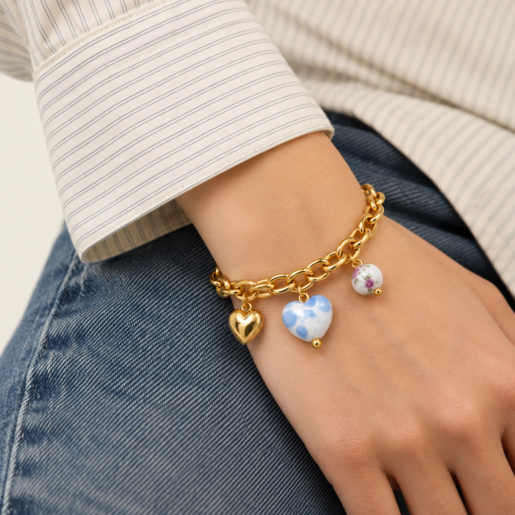 A close-up of a person's wrist wearing a gold chain bracelet with three charms: a gold heart, a light blue and white marbled heart, and a small white bead with purple floral details. The person is wearing a cream and blue striped shirt and blue jeans.