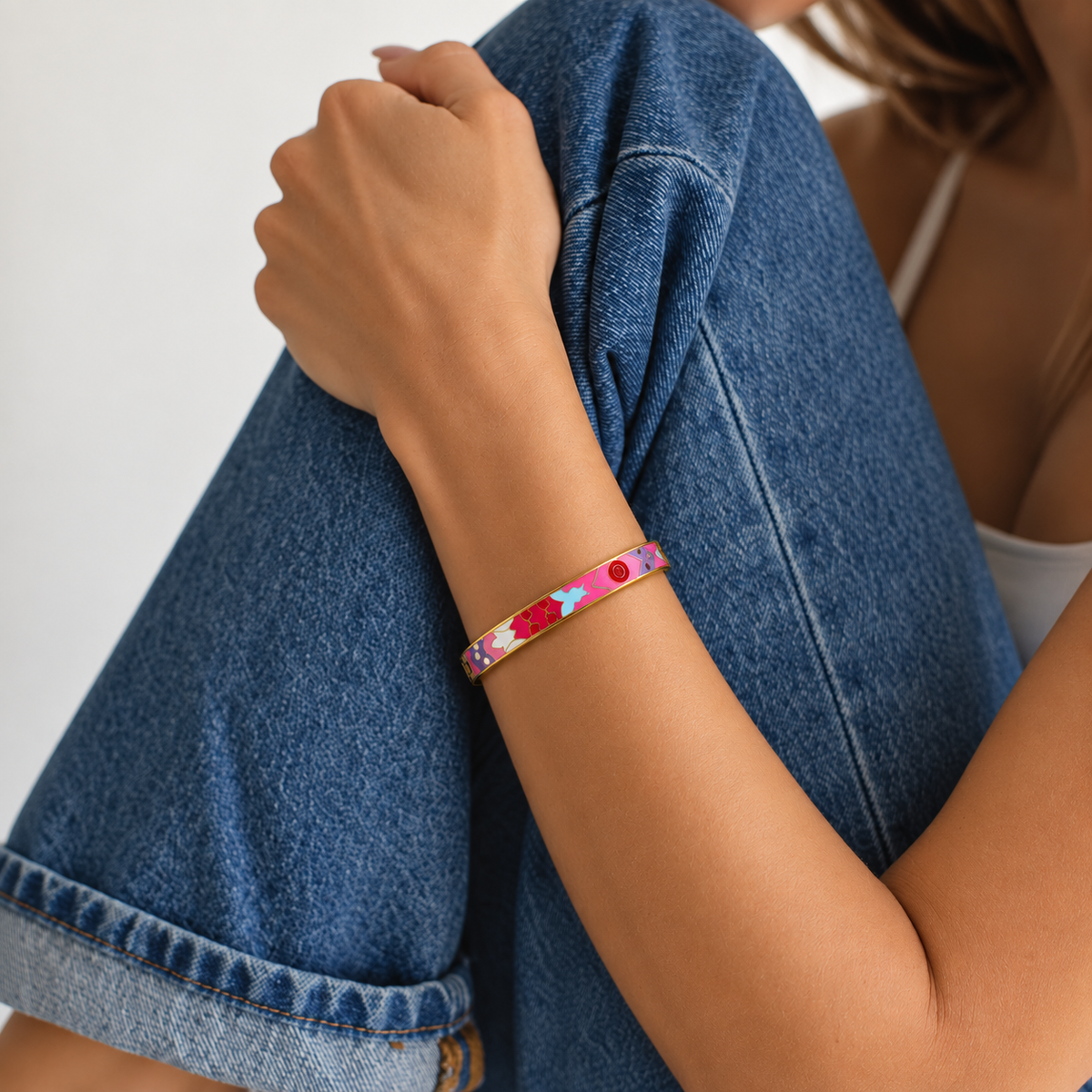A close-up of a person wearing a colorful enamel bangle bracelet on their wrist, with their arm resting on their knee in a blue denim jacket against a plain light background.