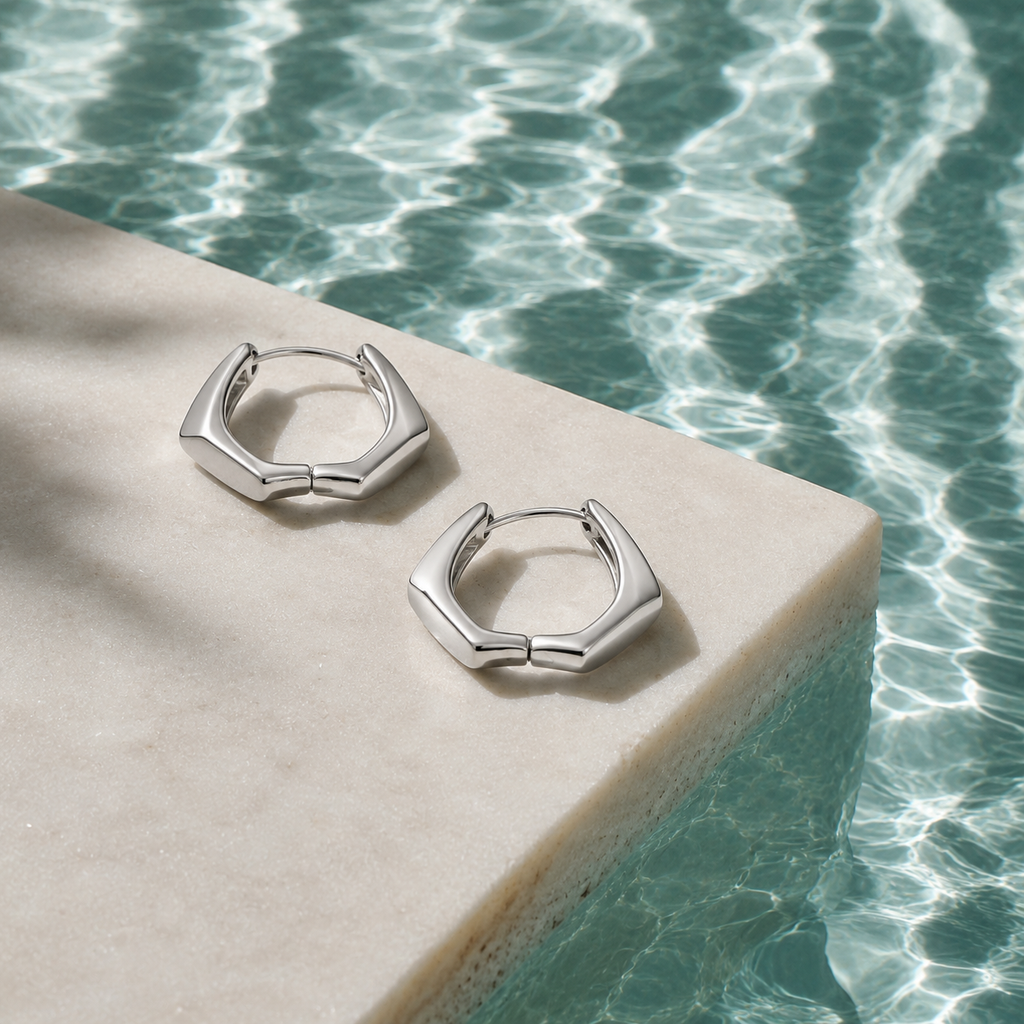 A pair of silver-toned geometric hoop earrings resting on a light beige stone slab beside rippling turquoise water.