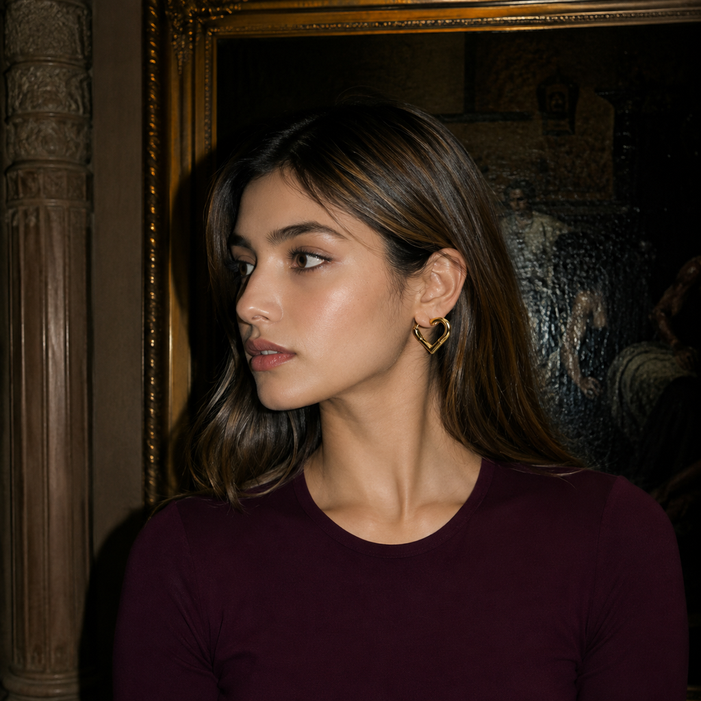 A woman with brown hair wearing a dark burgundy long-sleeve top and a gold heart-shaped earring, photographed in a dimly lit, elegant interior setting.