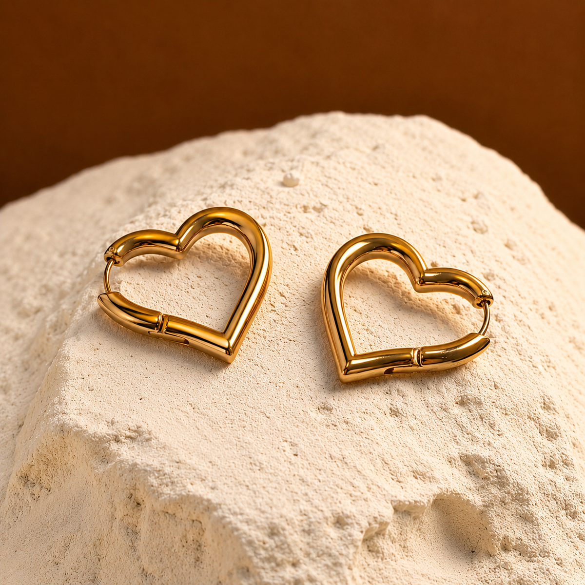 A pair of gold heart-shaped hoop earrings resting on a mound of textured beige powder against a warm brown background.
