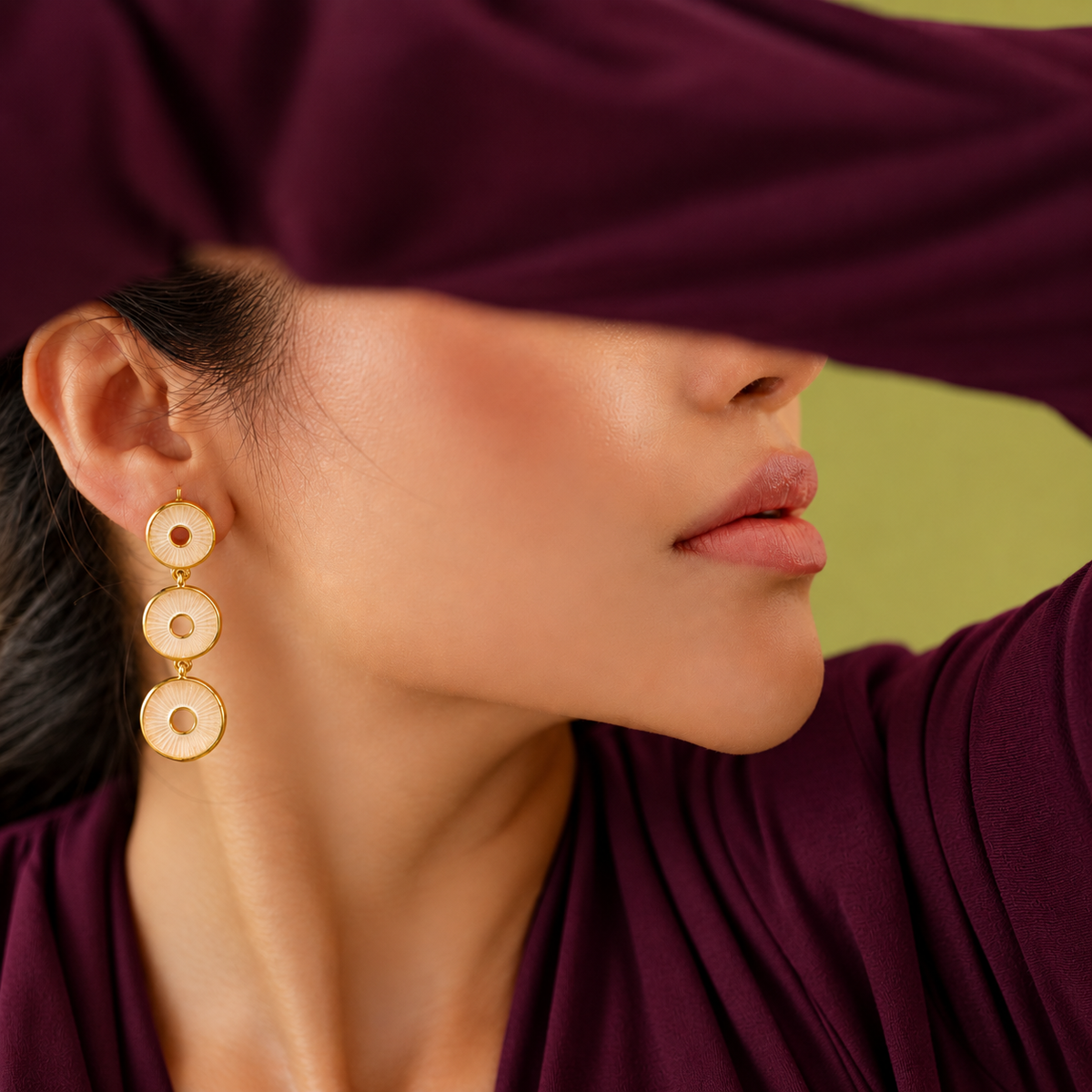 A close-up lifestyle shot of a model wearing gold drop earrings with circular disc charms, styled with a deep purple top and a matching purple blindfold against a soft olive green background.
