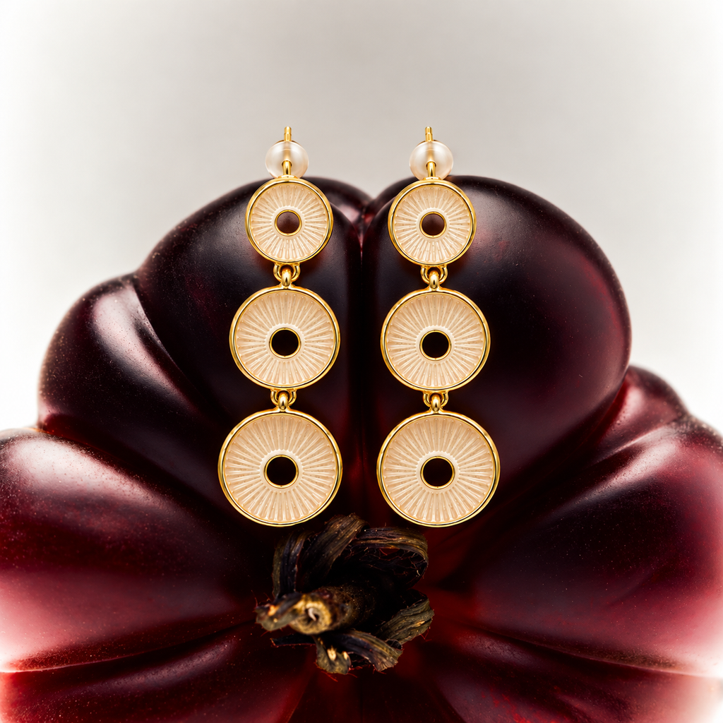 A pair of gold drop earrings with three circular disc charms on each earring, displayed on a dark red flower against a light gray background.