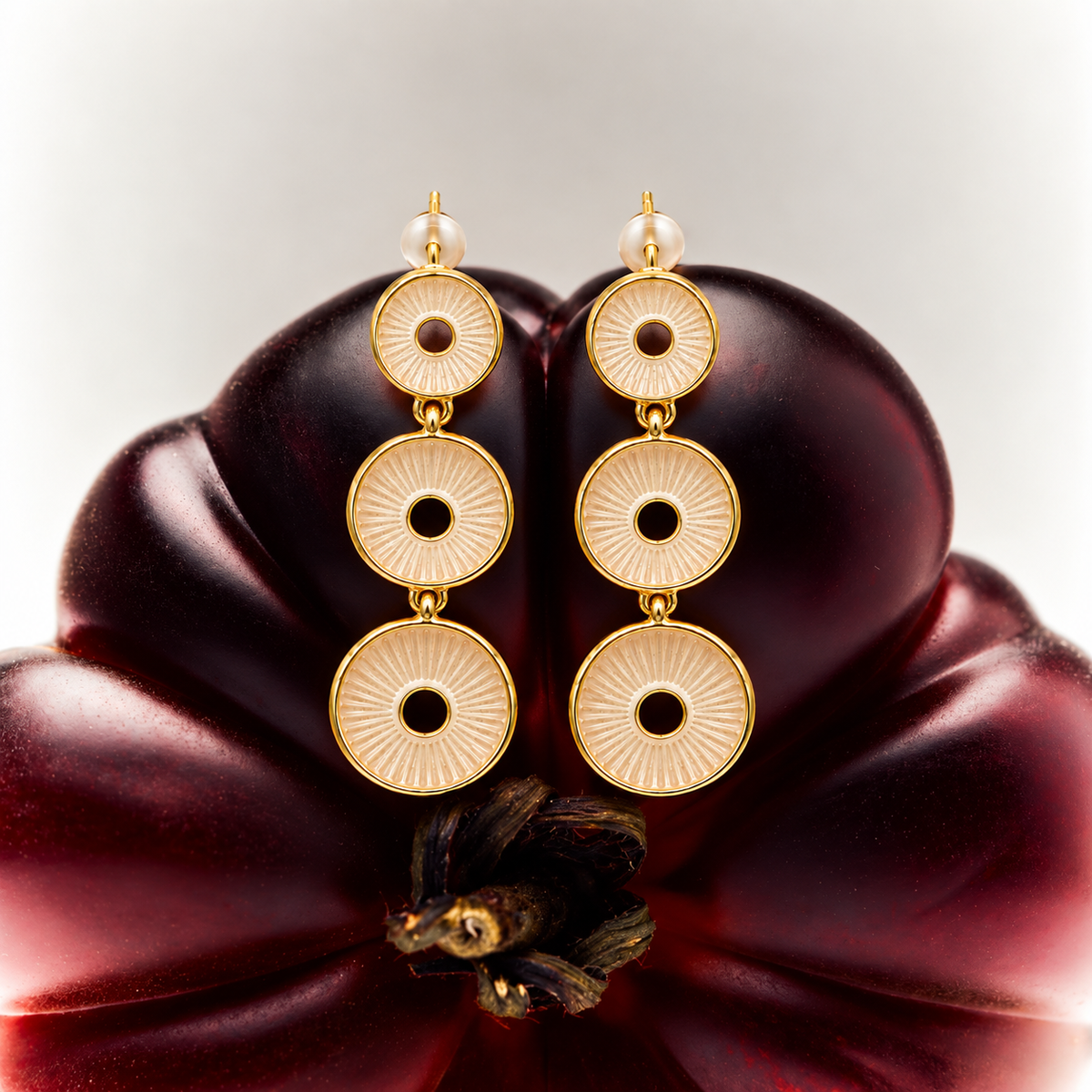 A pair of gold drop earrings with three circular disc charms on each earring, displayed on a dark red flower against a light gray background.