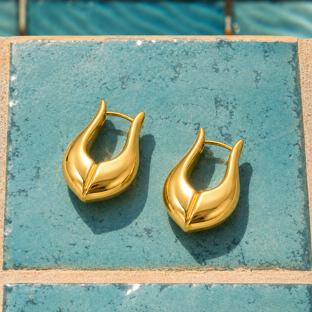 A pair of polished gold huggie hoop earrings with a leaf-like curved design, resting on a textured teal tile surface.