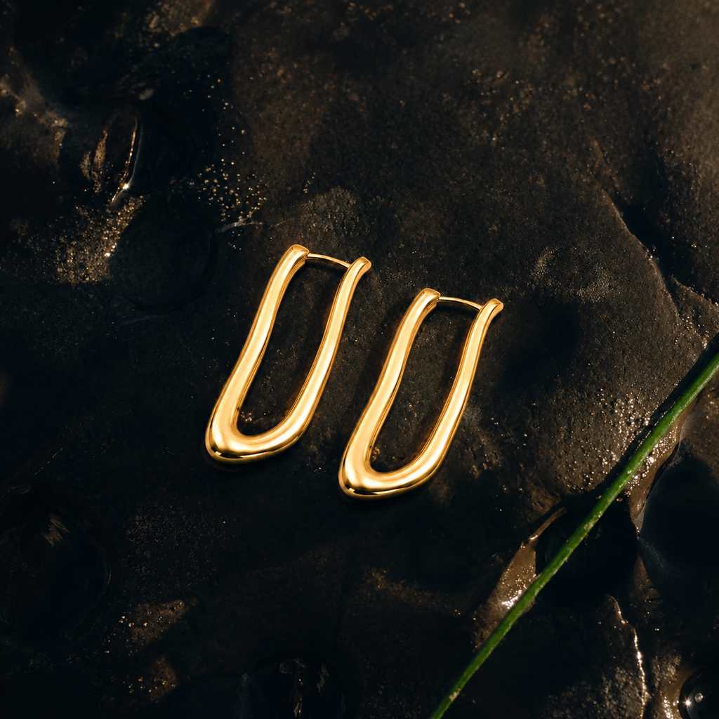 A pair of gold-toned, abstract curved hoop earrings resting on a dark, textured surface with water droplets.