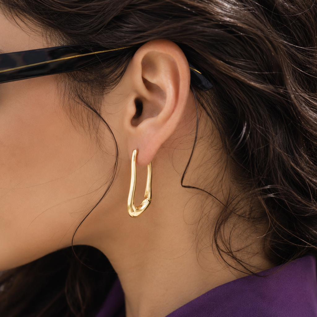 A close-up of a person's ear wearing a polished gold huggie hoop earring, with dark hair and part of a purple garment visible.