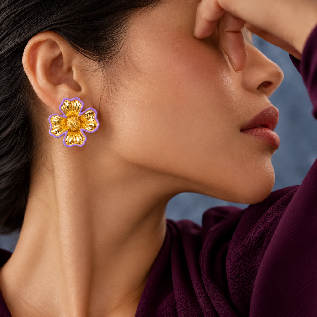 A close-up of a model wearing a gold flower-shaped stud earring with a purple outer border, set against a soft blue-gray background.