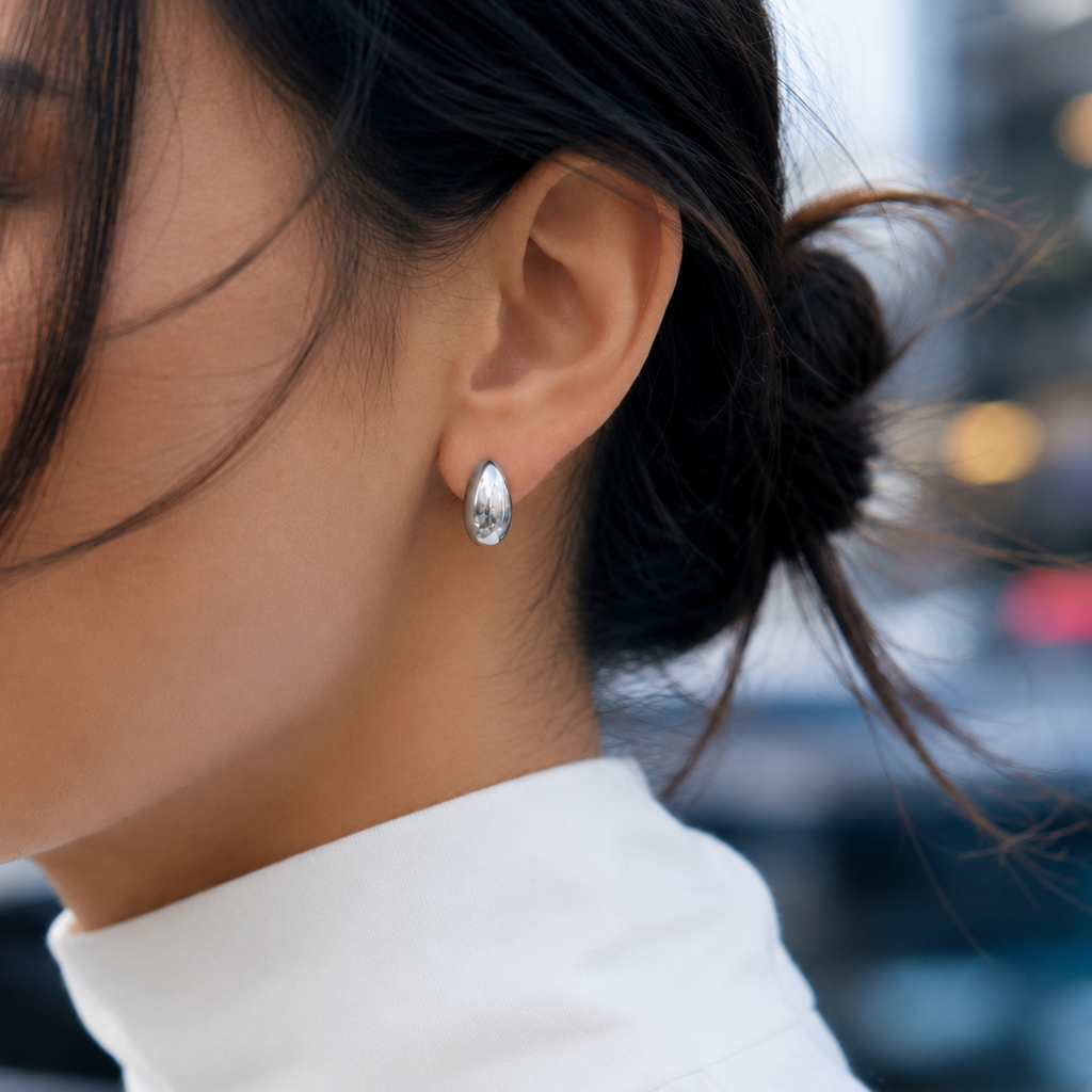 A close-up of a woman's ear wearing a small silver teardrop-shaped stud earring with a clear faceted gemstone, set against a softly blurred outdoor background.