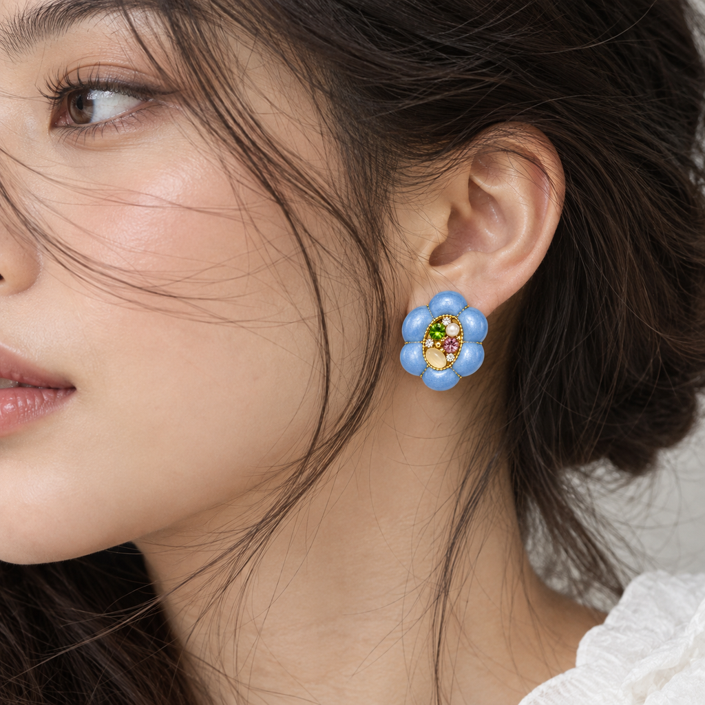 A close-up of a woman's ear wearing a flower-shaped stud earring with light blue petals, a gold center, and small multicolored gemstones.