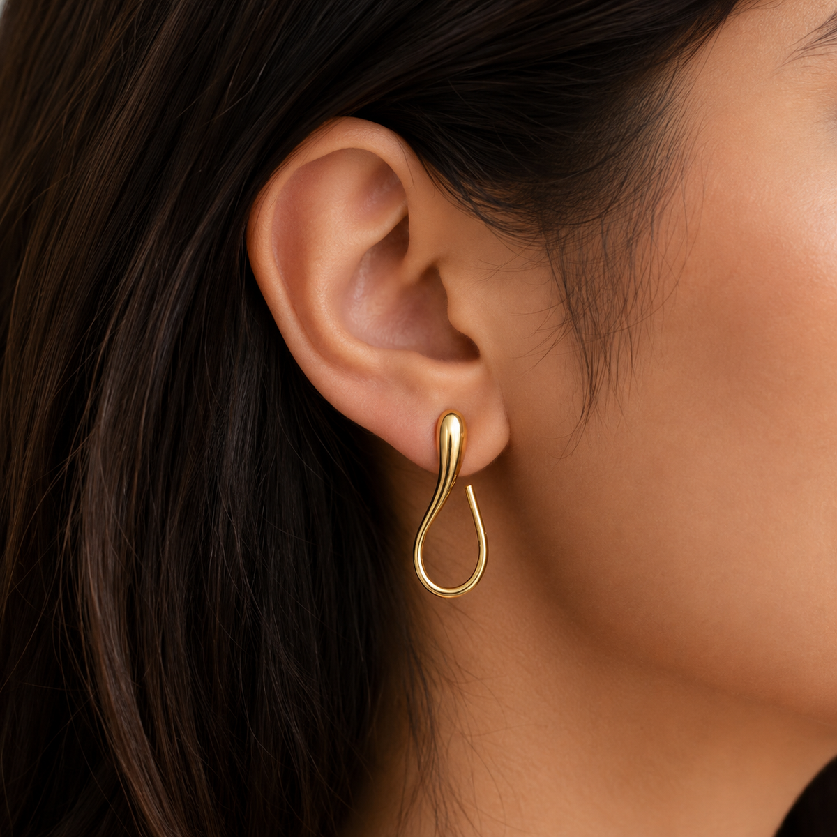 A close-up of a model's ear wearing a polished gold teardrop-shaped hoop earring.