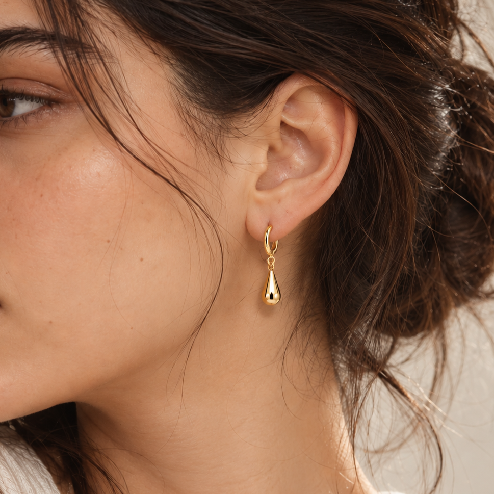 A close-up of a woman's ear wearing a small gold huggie hoop earring with a dangling teardrop charm.