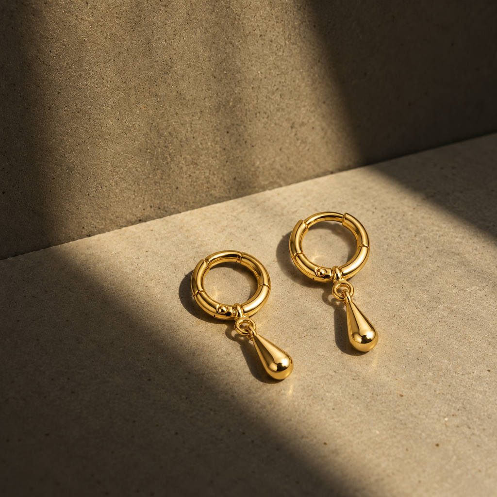 A pair of gold hoop earrings with teardrop dangles, displayed on a textured beige surface under dramatic, warm sunlight and shadow.