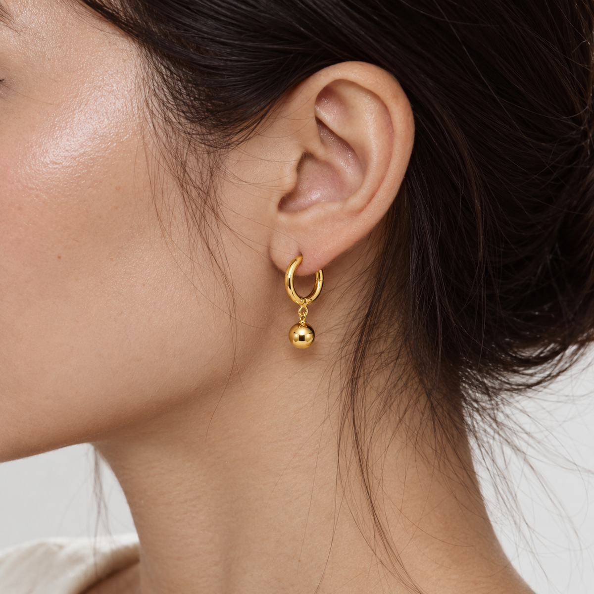 A close-up of a model's ear wearing a delicate gold huggie hoop earring with a small dangling gold bead.
