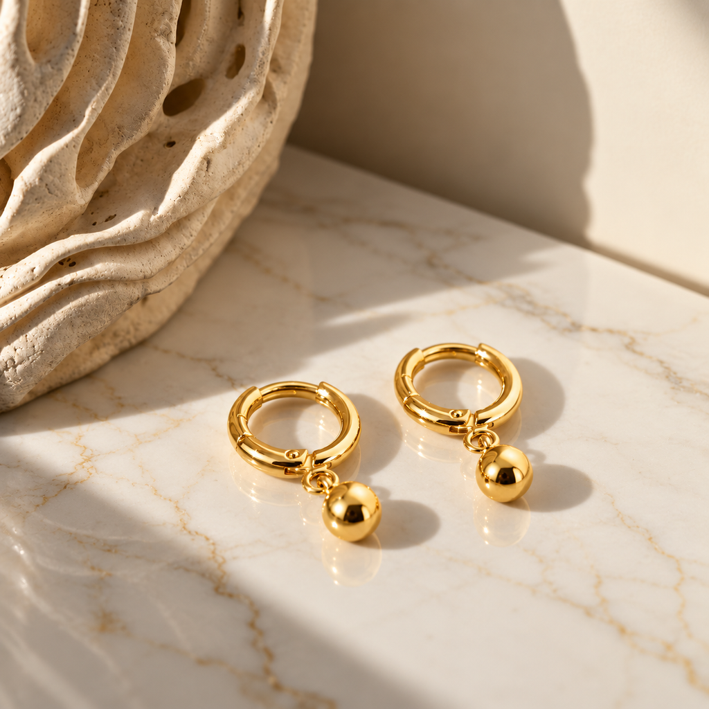 A pair of gold huggie hoop earrings with dangling polished ball charms, displayed on a white marble surface with a textured beige stone prop in the background.