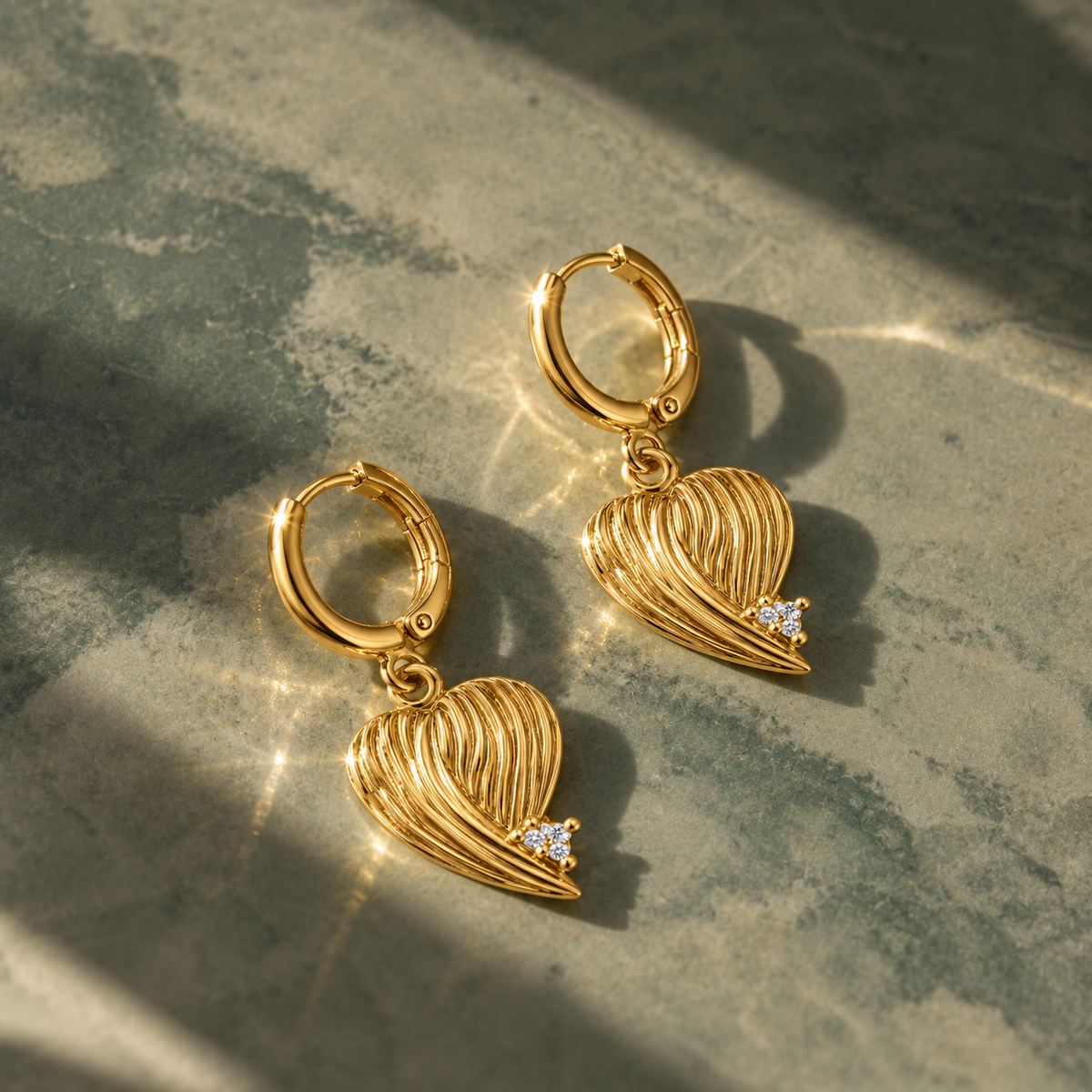 A pair of gold hoop earrings with textured heart-shaped dangles and small clear gemstone accents, displayed on a gray stone surface with dramatic sunlight and shadows.