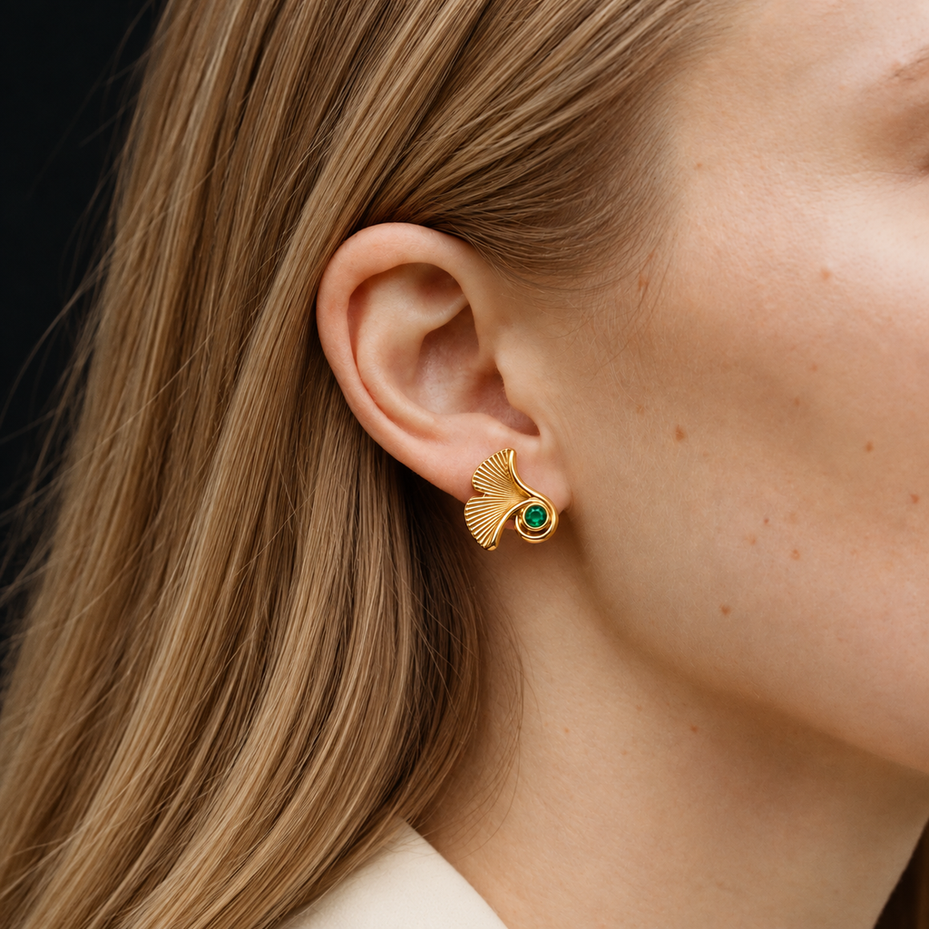A close-up of a woman's ear wearing a gold ginkgo leaf stud earring with a small green stone at the center, set against her hair and skin.
