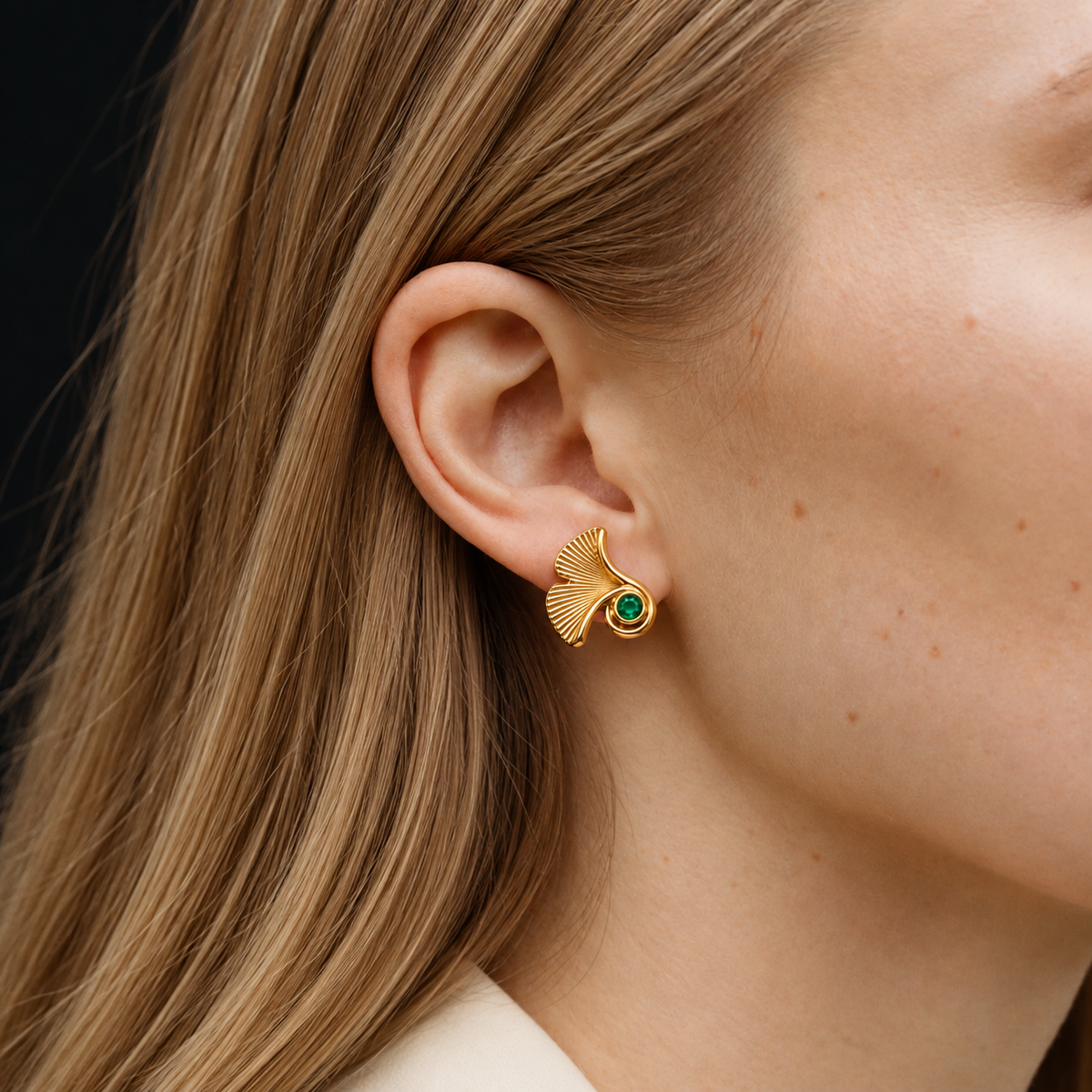 A close-up of a woman's ear wearing a gold ginkgo leaf stud earring with a small green stone at the center, set against her hair and skin.