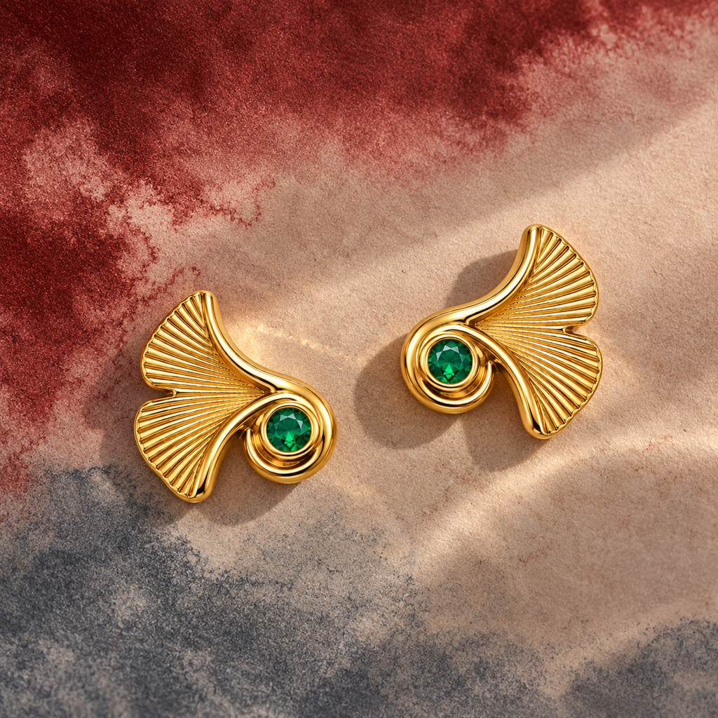A pair of gold-toned statement earrings with a fan-shaped leaf design and a round green gemstone, displayed on a textured beige surface with a marbled red and gray background.
