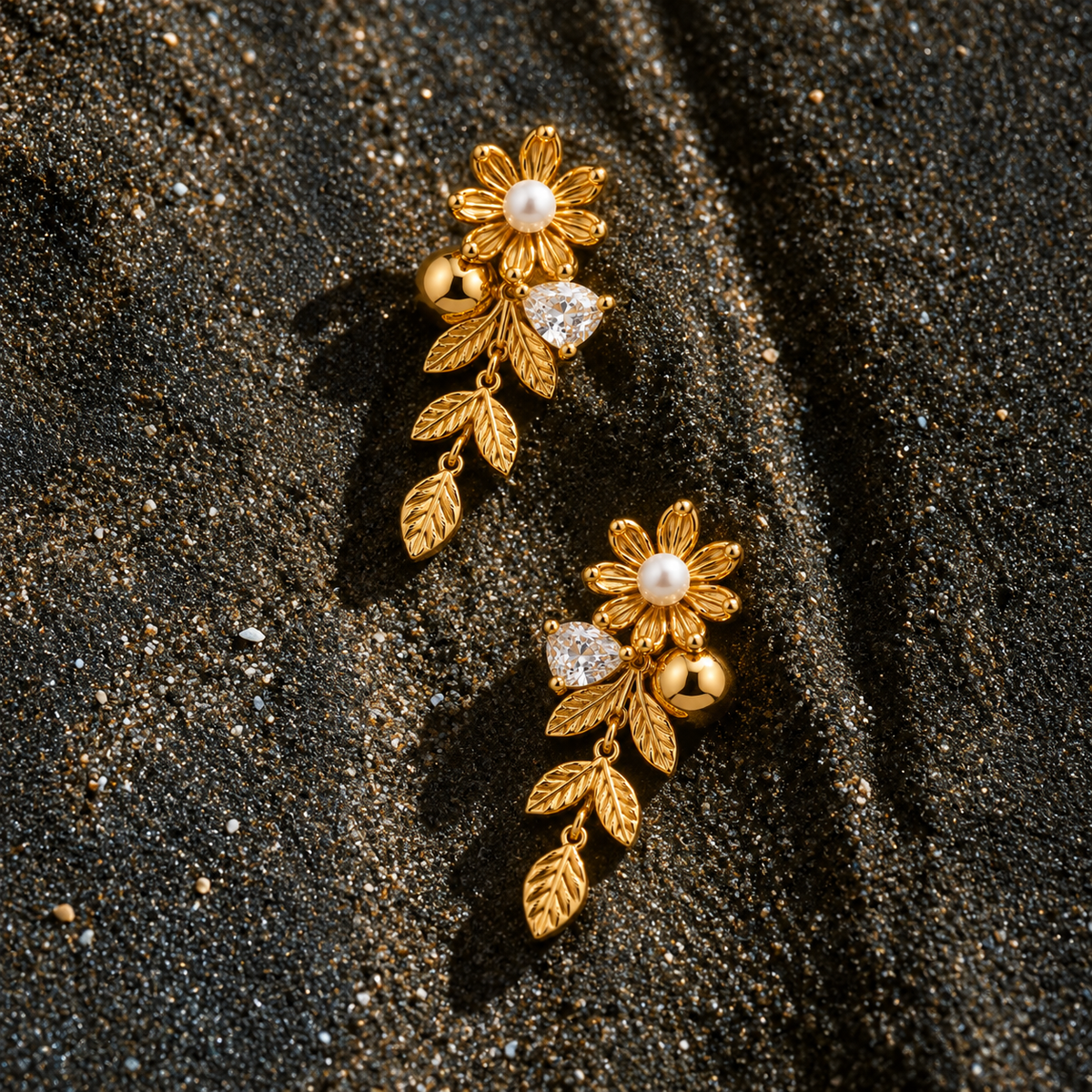 A pair of gold floral drop earrings with pearl centers, clear heart-shaped stones, and leaf charms, resting on dark textured sand.
