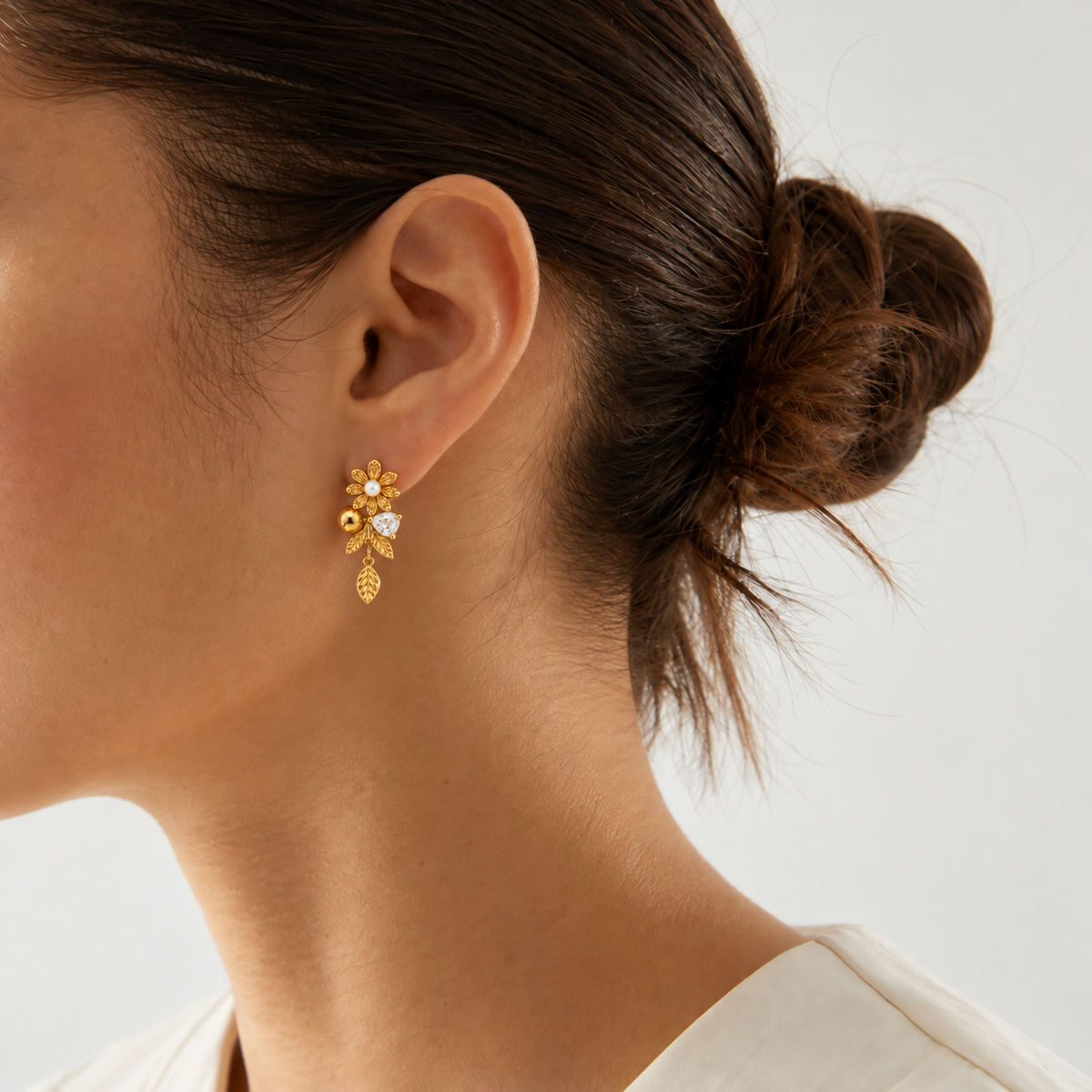 A close-up side profile of a woman wearing a delicate gold dangle earring with a small floral cluster and a clear heart-shaped stone, set against a clean light gray background.