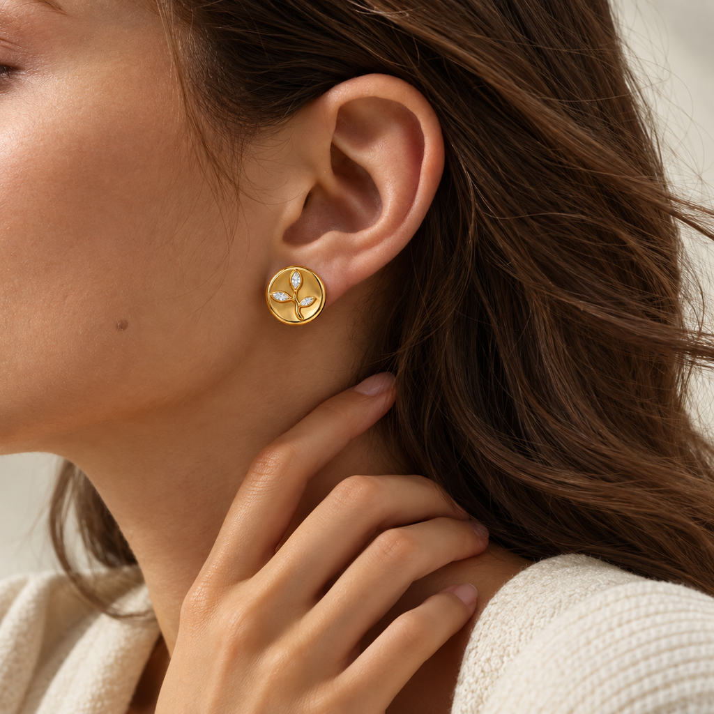 A close-up of a woman wearing a small gold stud earring with a white floral or clover-like motif, against a soft light background.