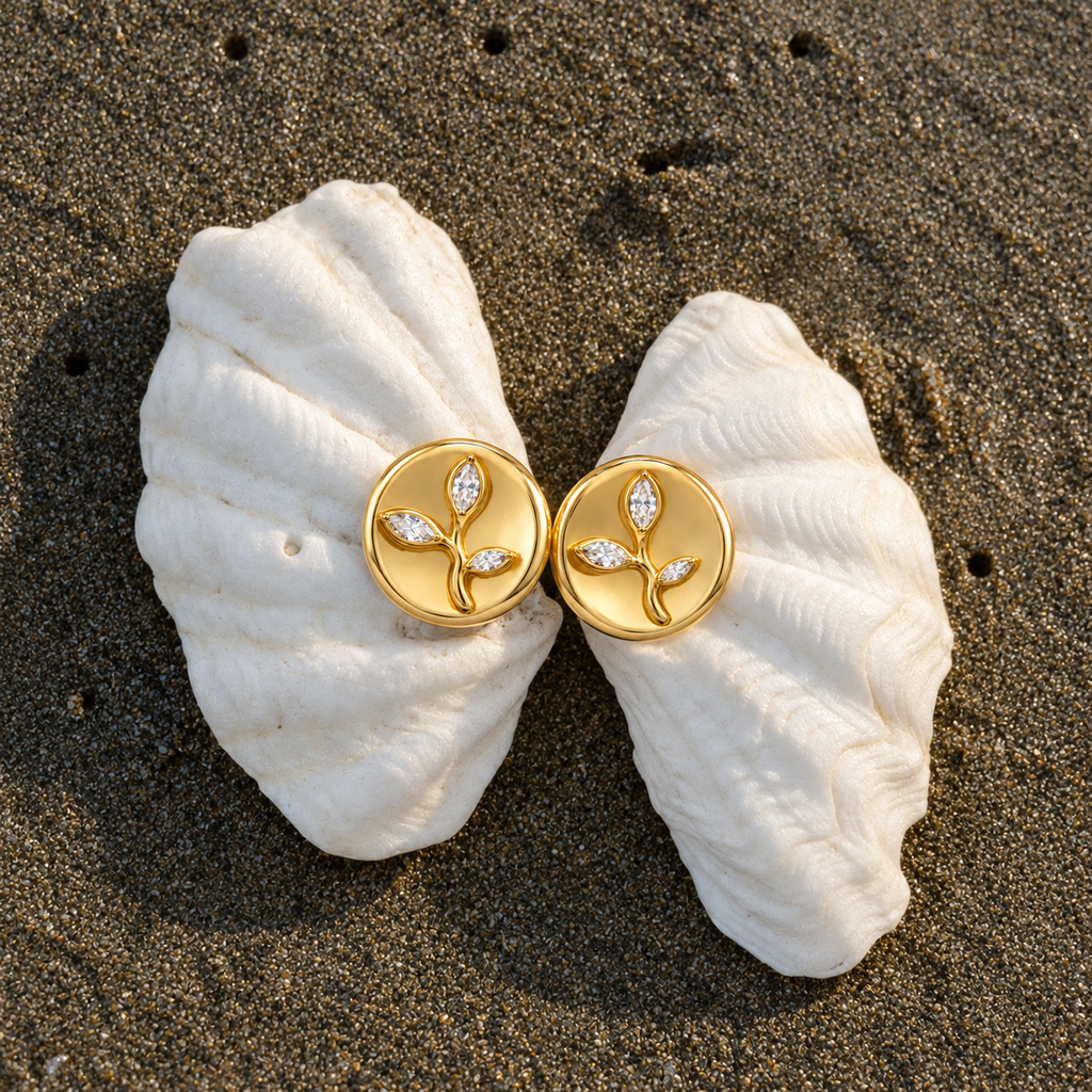A pair of gold leaf stud earrings with small clear crystal accents, displayed on a white clam shell over dark sand.