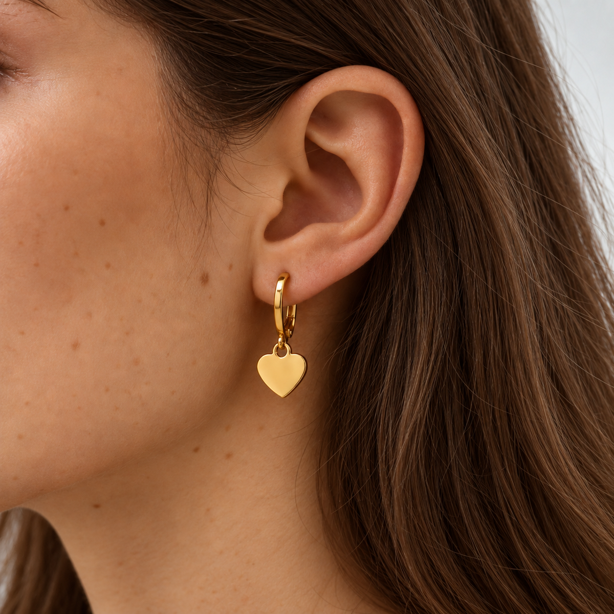 A close-up of a woman's ear wearing a small gold huggie hoop earring with a dangling polished heart charm.
