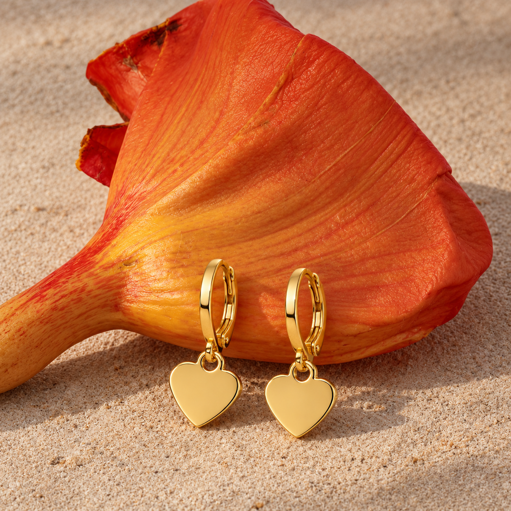 A pair of gold hoop earrings with dangling heart charms, displayed on a sandy surface next to a large orange flower.