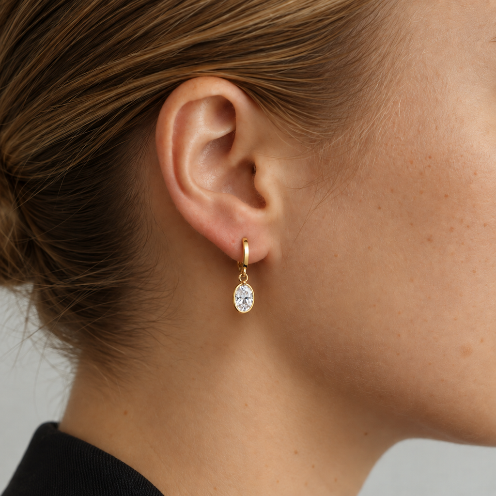 A close-up of a model's ear wearing a delicate gold huggie hoop earring with a small oval clear gemstone.