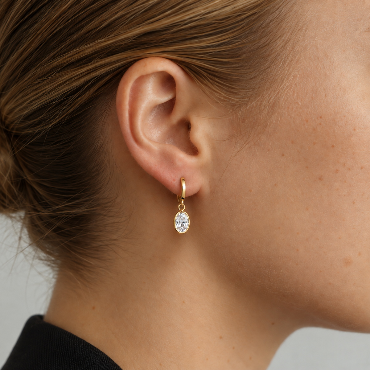 A close-up of a model's ear wearing a delicate gold huggie hoop earring with a small oval clear gemstone.