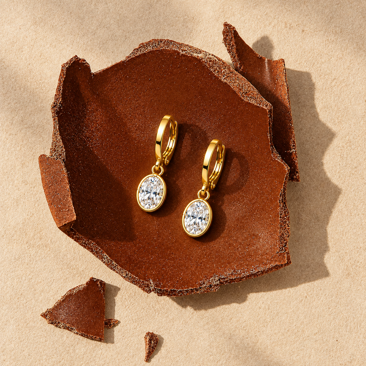 A pair of gold hoop earrings with oval clear gemstones, displayed on a textured brown ceramic dish over a sandy beige background.