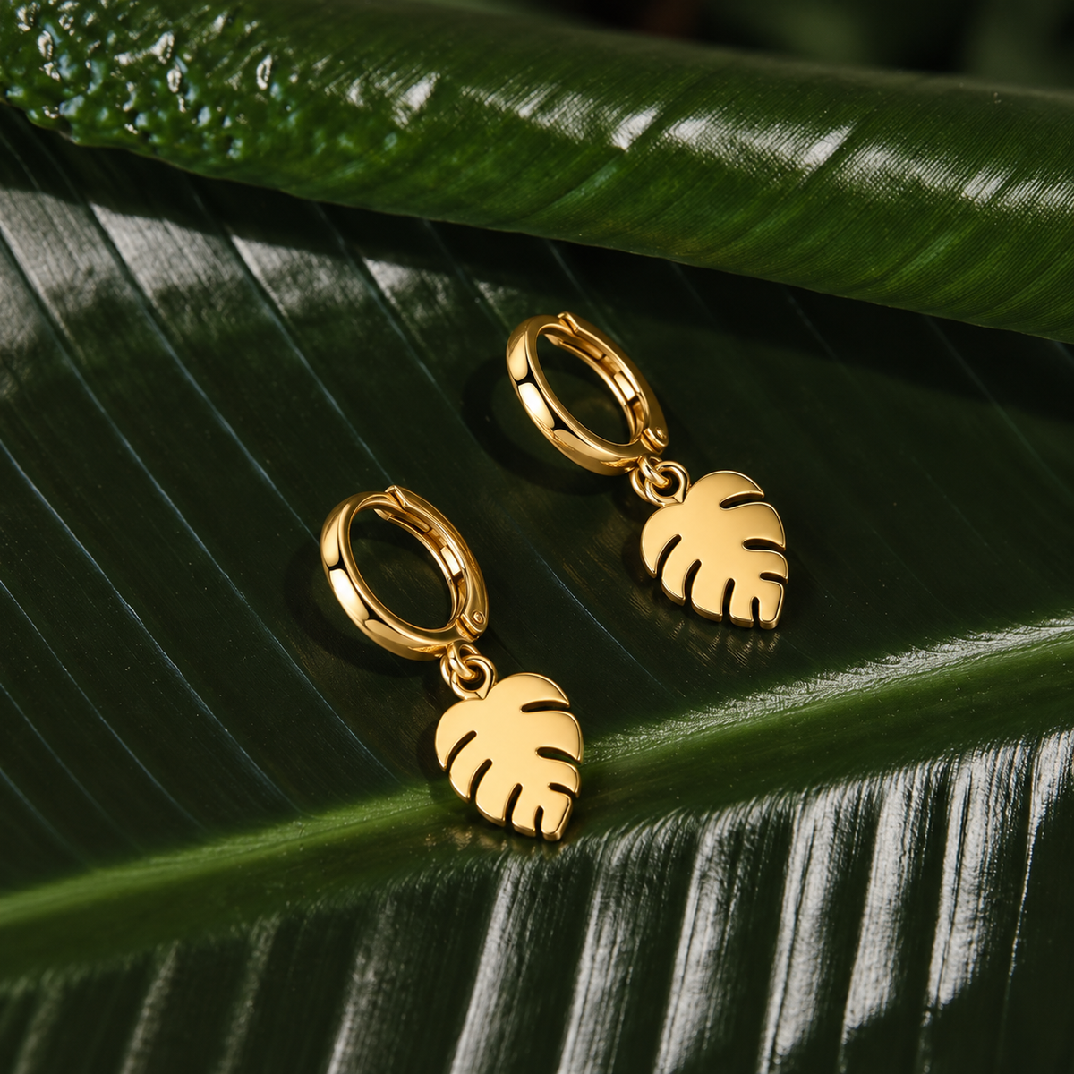 A pair of gold hoop earrings with dangling monstera leaf charms, displayed on a large green leaf.