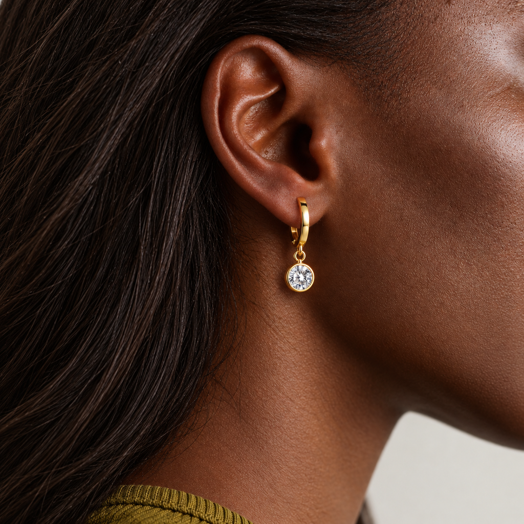 A close-up of a model's ear wearing a small gold huggie hoop earring with a clear round gemstone dangle.