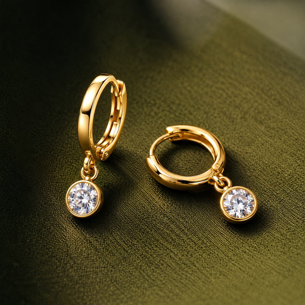 A pair of gold huggie hoop earrings, each with a small round bezel-set clear gemstone dangle, displayed on a dark olive green textured surface.