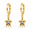 A pair of gold huggie hoop earrings with dangling turtle charms. The turtle charms feature blue sapphires set in a halo of clear pavé stones, shown on a clean white background.