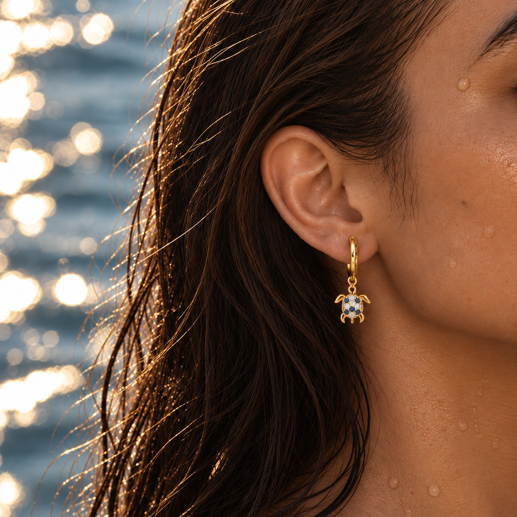 A close-up lifestyle shot of a woman's ear wearing a gold huggie hoop earring with a small sea turtle charm. The charm features white enamel inlay and tiny blue stones.