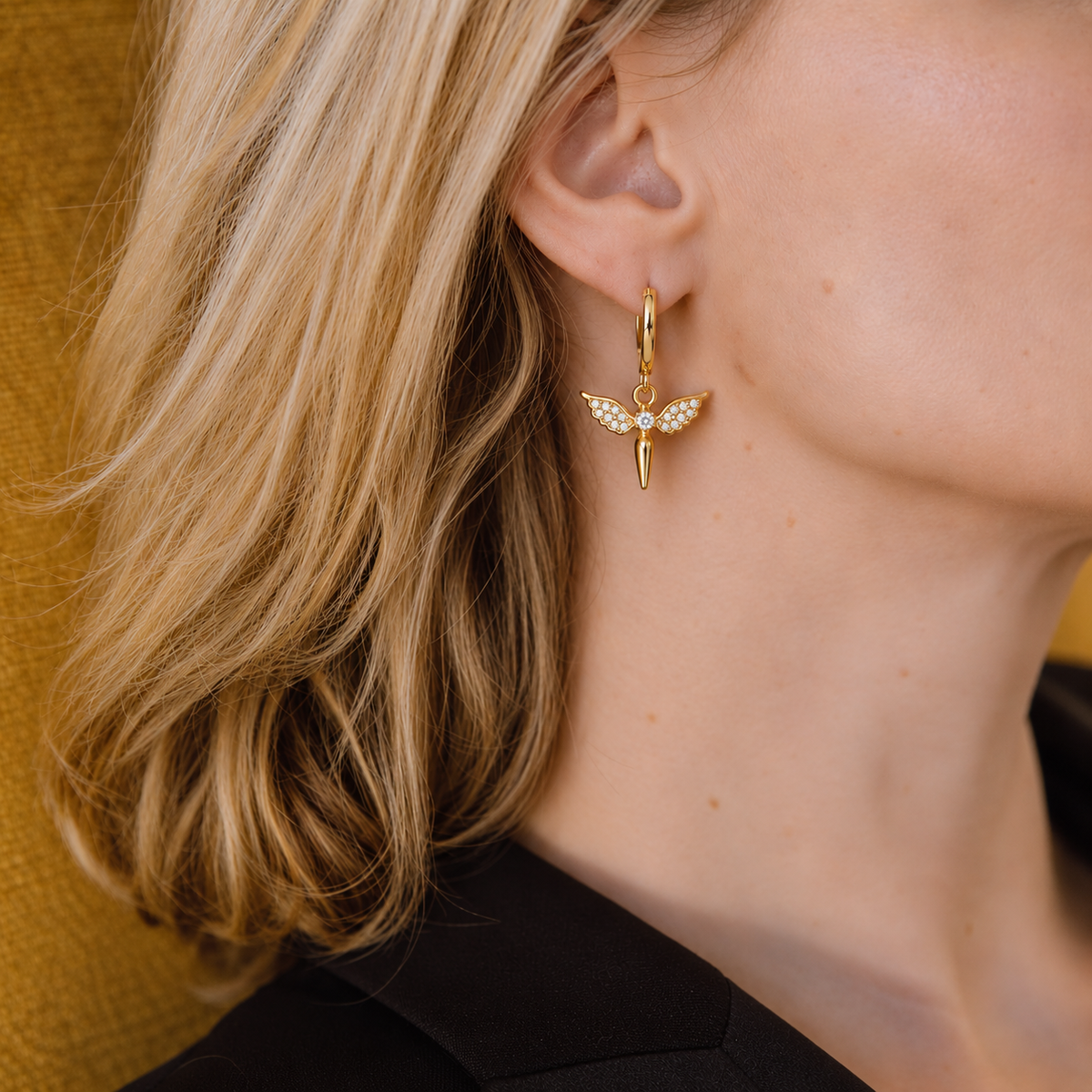 A close-up lifestyle shot of a woman wearing a delicate gold huggie hoop earring with a winged fairy charm adorned with small white crystals.