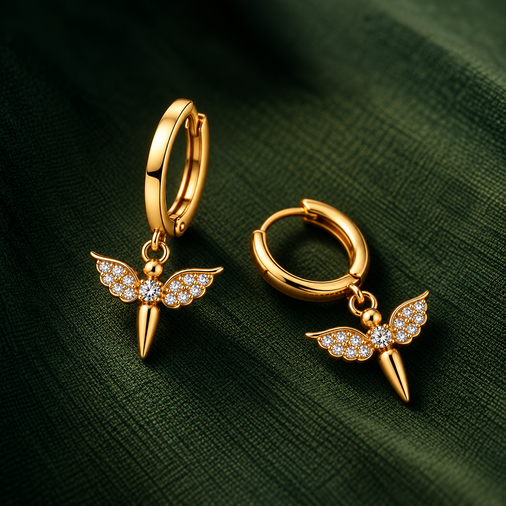 A pair of gold huggie hoop earrings with wing-shaped dangles adorned with small clear crystals, displayed on a dark green textured fabric background.