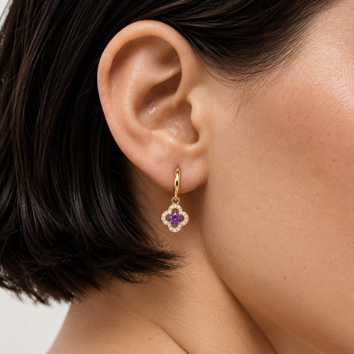 A close-up of a model's ear wearing a delicate gold dangle earring with a purple flower-shaped center and small clear stones around it.