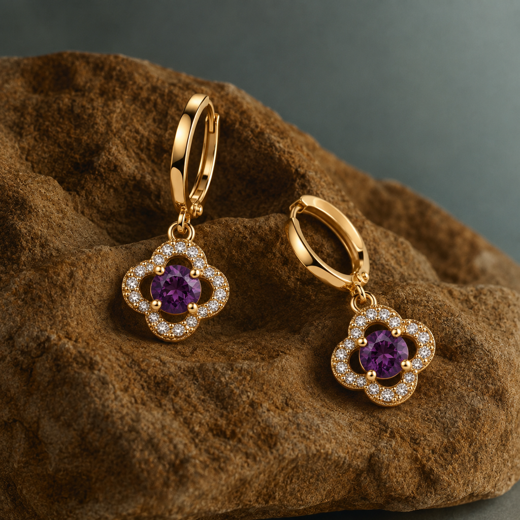 A pair of gold hoop earrings with flower-shaped charms featuring purple gemstones and clear accent stones, displayed on a textured brown rock against a dark gray background.