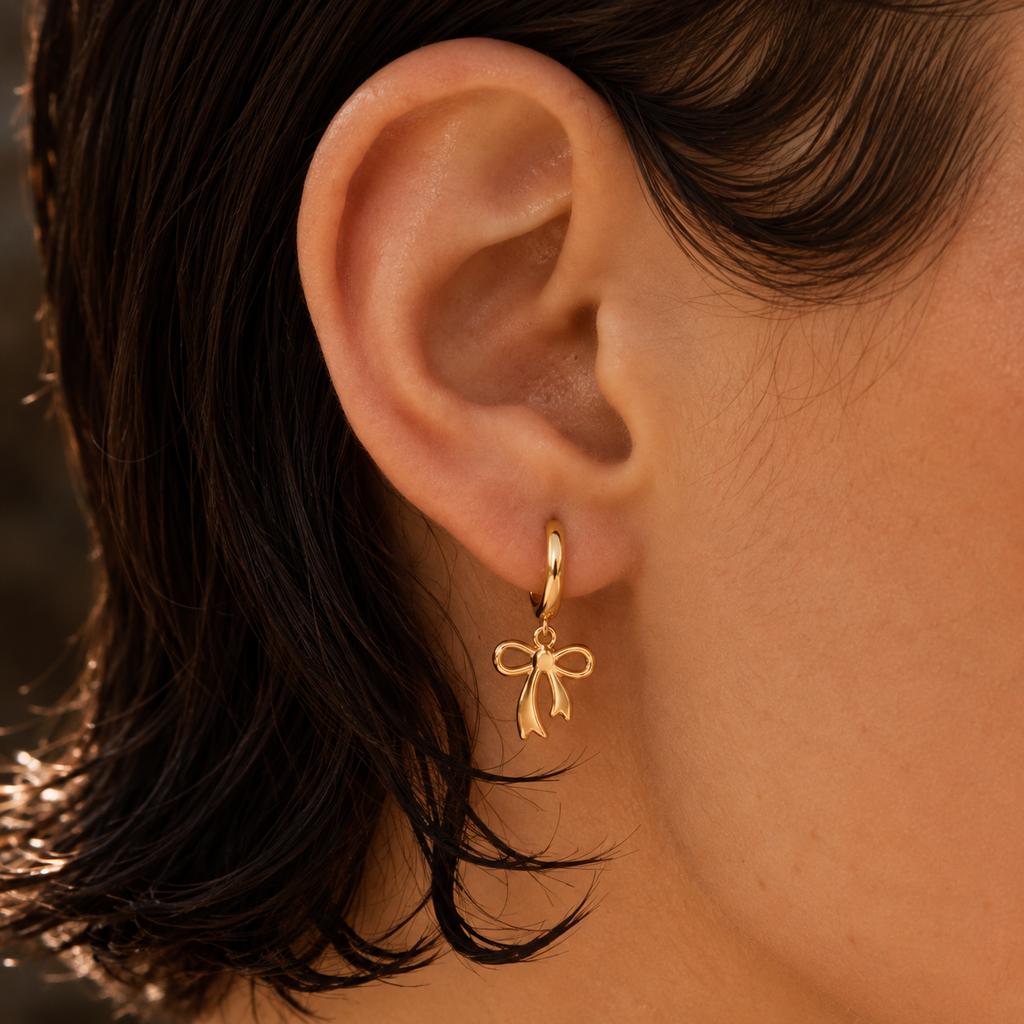 A close-up of a person's ear wearing a delicate gold huggie hoop earring with a small bow charm, set against dark hair and warm skin tones.