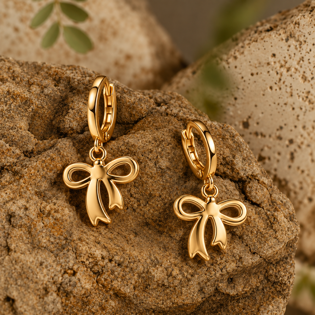 A pair of gold hoop earrings with dangling bow charms, displayed on textured brown rocks with a soft-focus natural background.