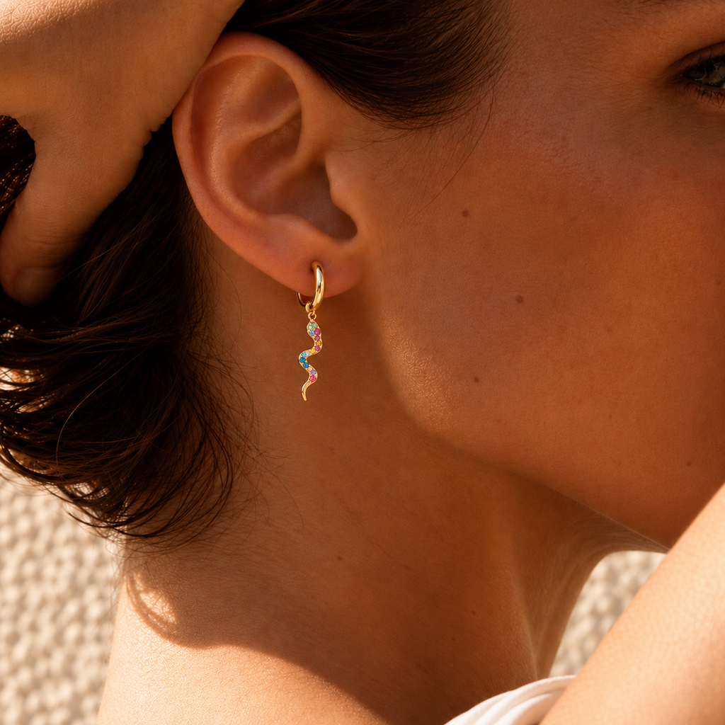 A close-up of a model's ear wearing a delicate gold huggie hoop earring with a small wavy drop charm adorned with pastel rainbow-colored stones.