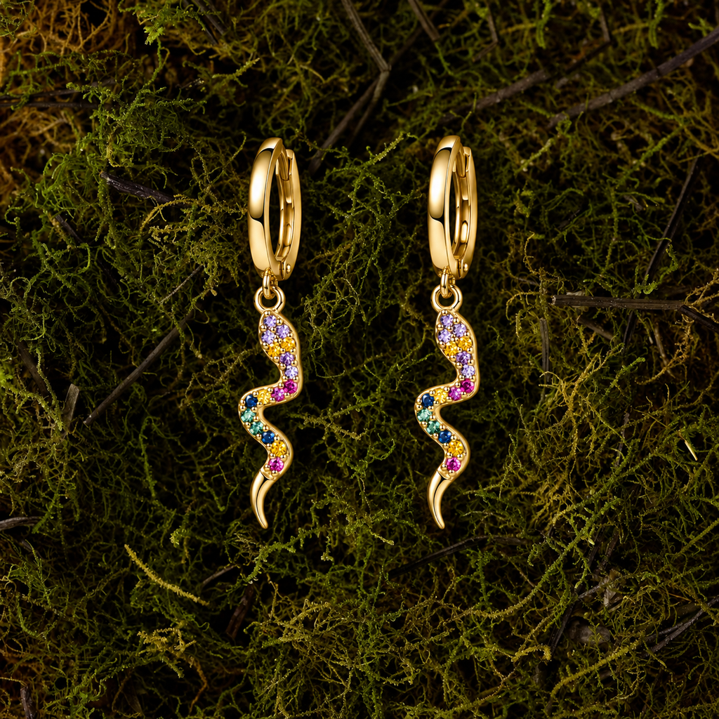 A pair of gold hoop dangle earrings with a wavy, snake-like charm set with small multicolored gemstones, displayed on a bed of green moss.