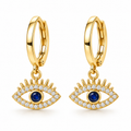 A pair of gold huggie hoop earrings with dangling evil eye charms. The charms feature a gold border paved with small clear cubic zirconia stones and a central round dark blue cabochon stone.