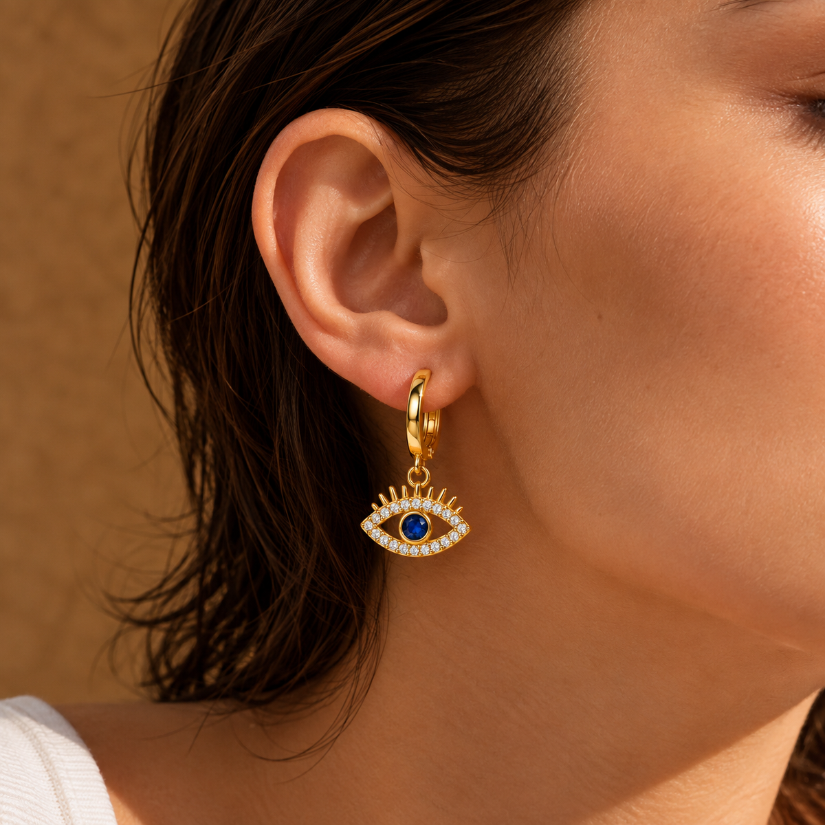 A close-up of a model's ear wearing a gold huggie hoop earring with a dangling evil eye charm. The charm features a blue center stone surrounded by small white stones and gold rays, set against a warm brown textured background.