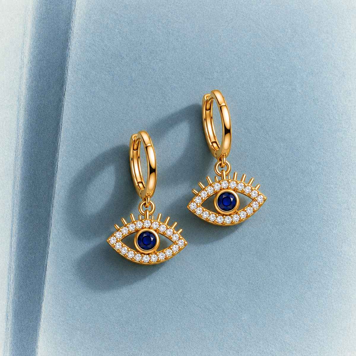 A pair of gold huggie hoop earrings with dangling evil eye charms. The charms feature a gold border paved with small clear stones, a deep blue circular center stone, and a matching blue center stone in the other earring.