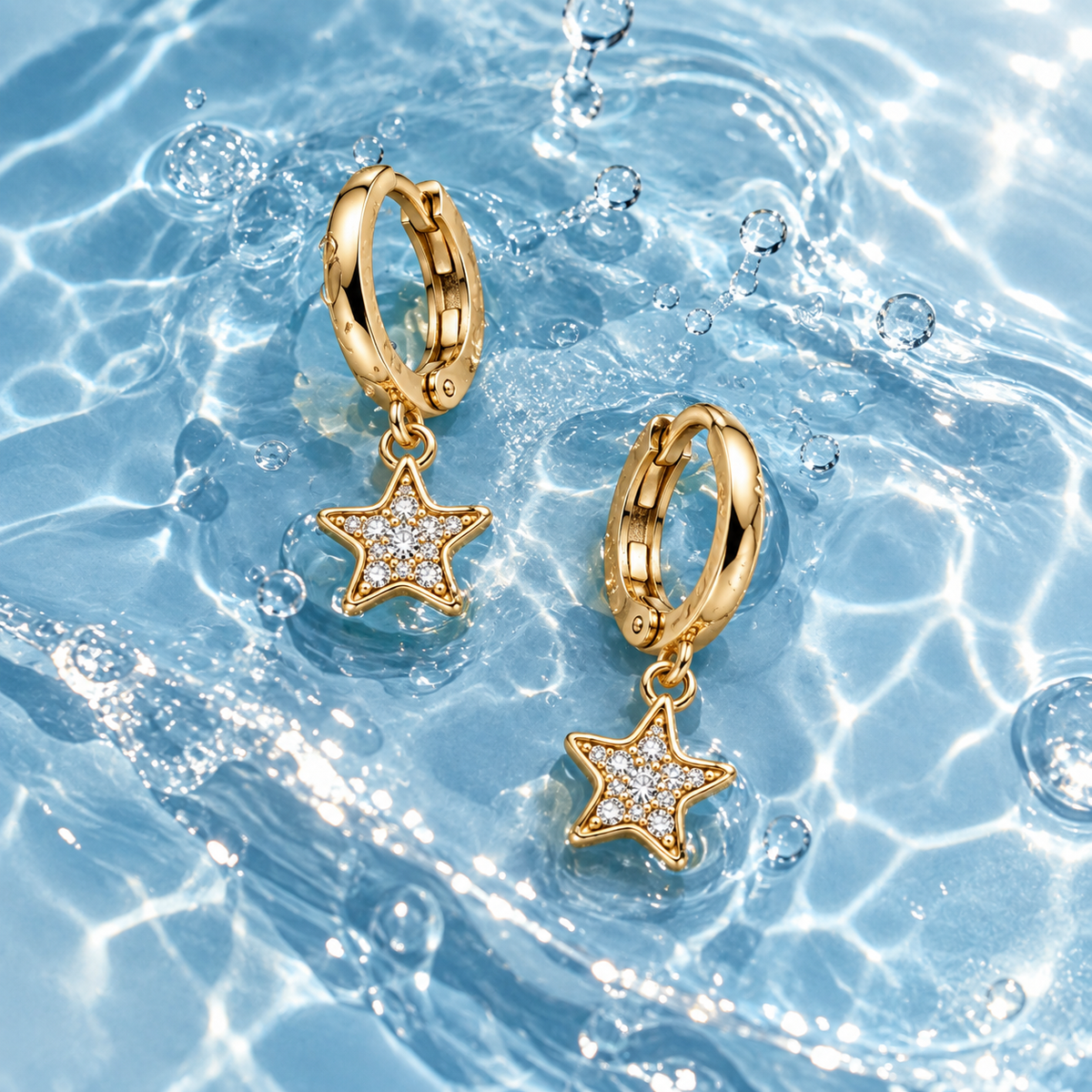 A pair of gold huggie hoop earrings with dangling star charms set with clear stones, resting on rippling blue water.